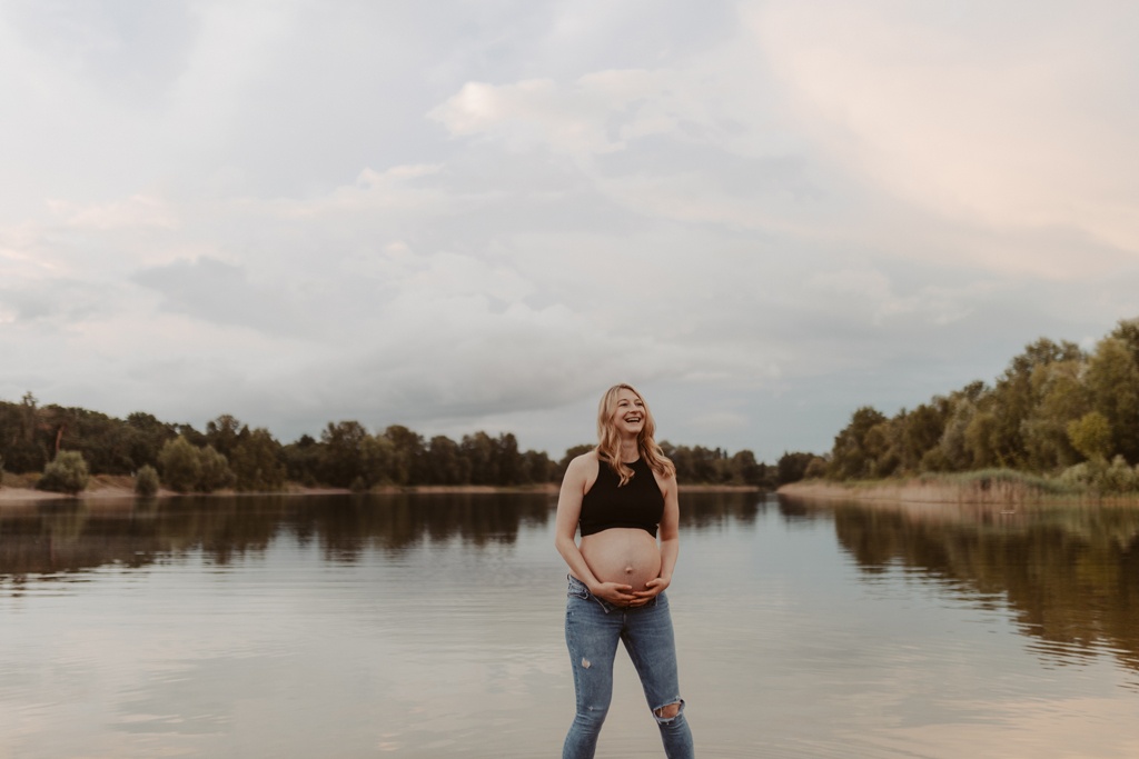 Babybauchfotos in berlin mit der Berliner Fotografin für Kinderfotografie Maria Brussig Lensofbeauty. fotos am See. Fotoshooting Babybauch im grünen. Wasser im Hintergrund. Paarfotos zusammen am See.